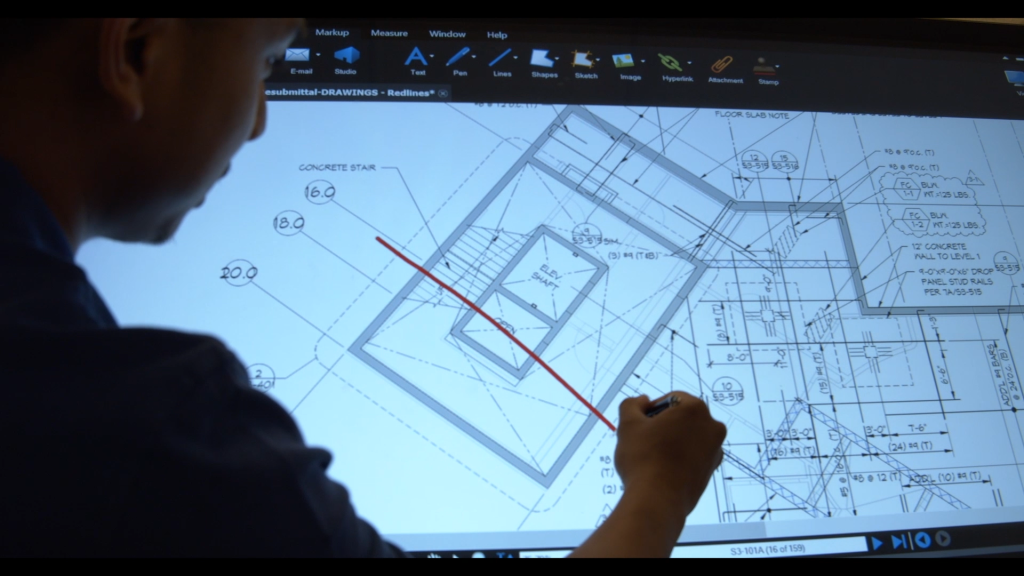 A person in a silhouette draws on a building plan on top of a lighted drafting table.