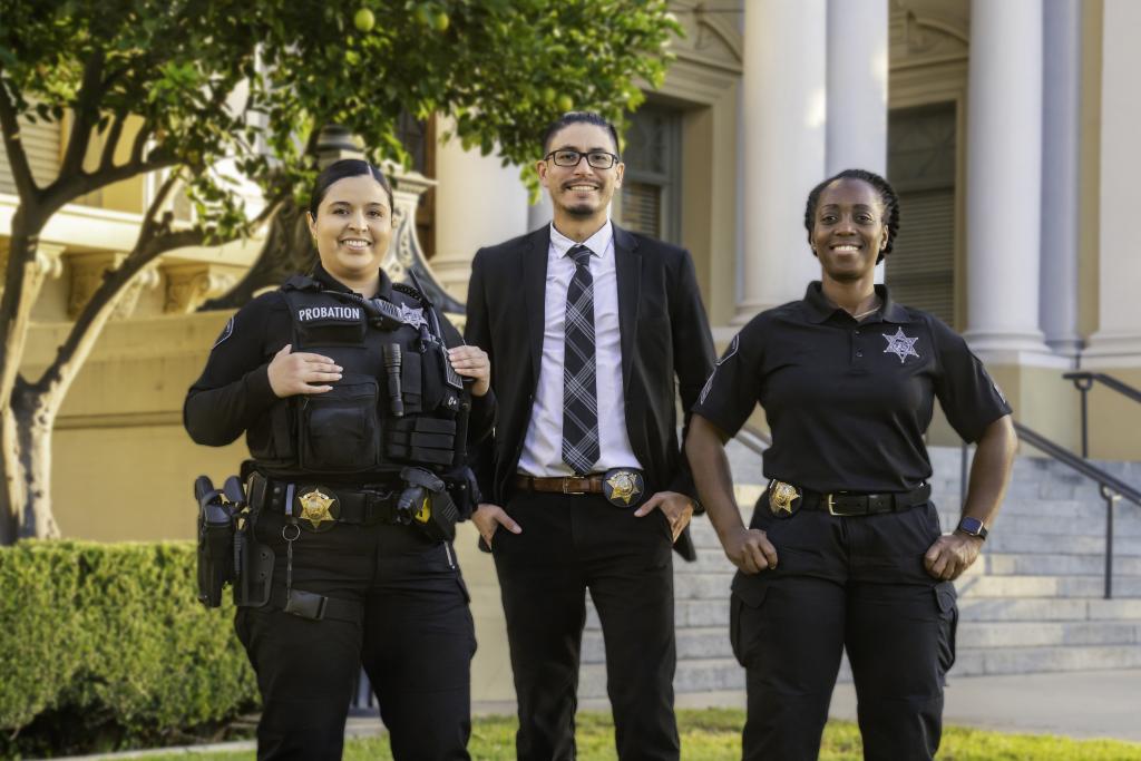 Three individuals stand in front of a courthouse. One wearing a law enforcement uniform, one in business attire and one in a black business casual shirt and pant uniform